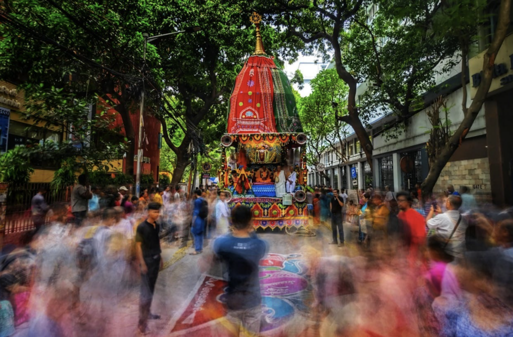 Ornate Ratha Yatra chariot surrounded by a blurred, colorful crowd in a tree-lined city street during a festival.
